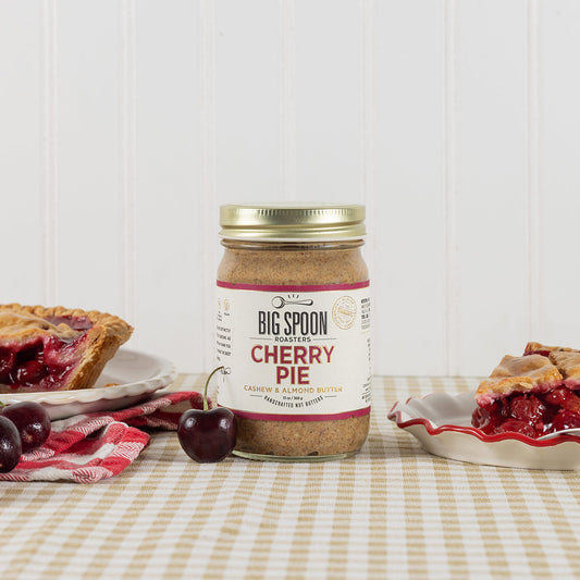One glass jar of nut butter on a gingham tablecloth surrounded by cherry pies and cherries