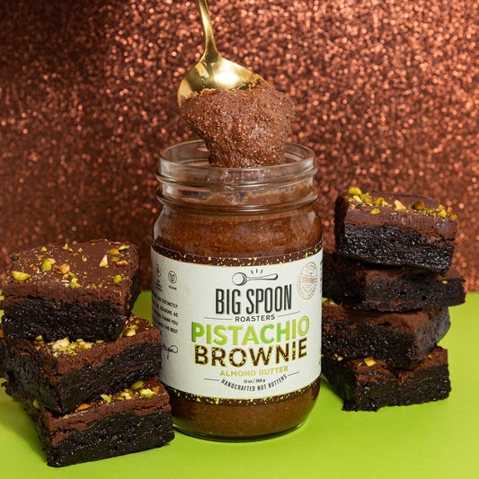 jar of pistachio brownie almond butter surrounded by pistachio brownies on a green and brown glitter background