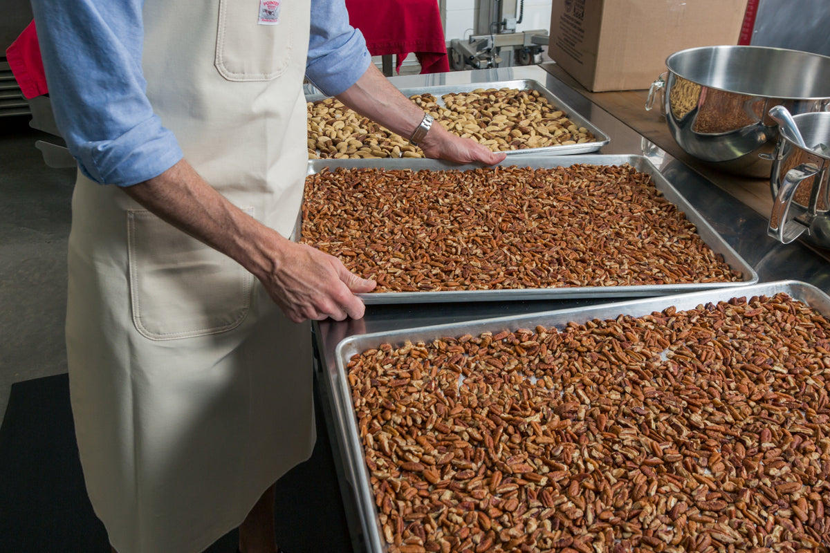 A man in a blue shirt and white apron holds a sheet pan of pecans with other sheet pans of nuts around it