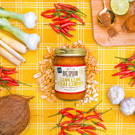 13oz Lum Lum Thai Curry Peanut & Cashew Butter surrounded by red chilis, peanuts, cashews, shallots, coconut and green onion. A cutting board topped with a golden spoon, coconut sugar, limes and ginger all sits on a yellow gingham cloth. 